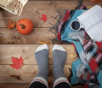 Person in warm socks with a book, hot drink, pumpkin, and blanket around them