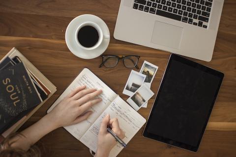 Picture shows a pair of hands writing in an open journal on a wooden brown table top. Next to the journal are a stack of books with the top being blue and titled Your Soul. On the table there is also a pair of black eyeglasses, a tablet, three polaroid pictures, a cup of coffee in a white cup on a white saucer, and an open laptop. 